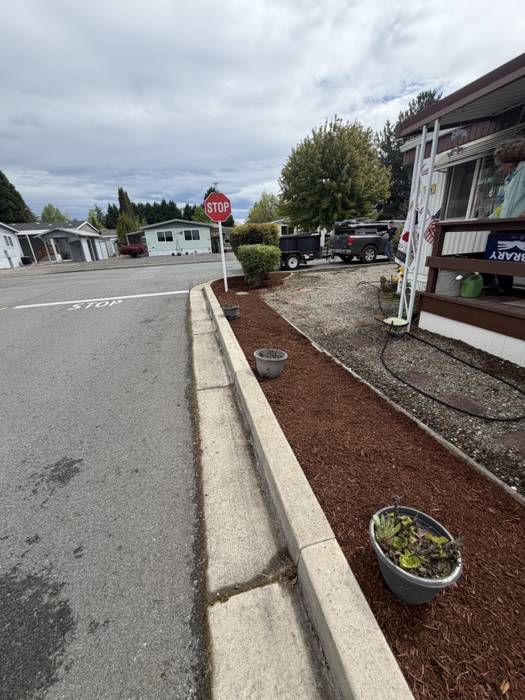 Fresh dark brown bark mulch spread in a narrow curbside planting bed beside a concrete curb at a Grants Pass, Oregon home, with potted succulents and a clean gravel strip along the porch.